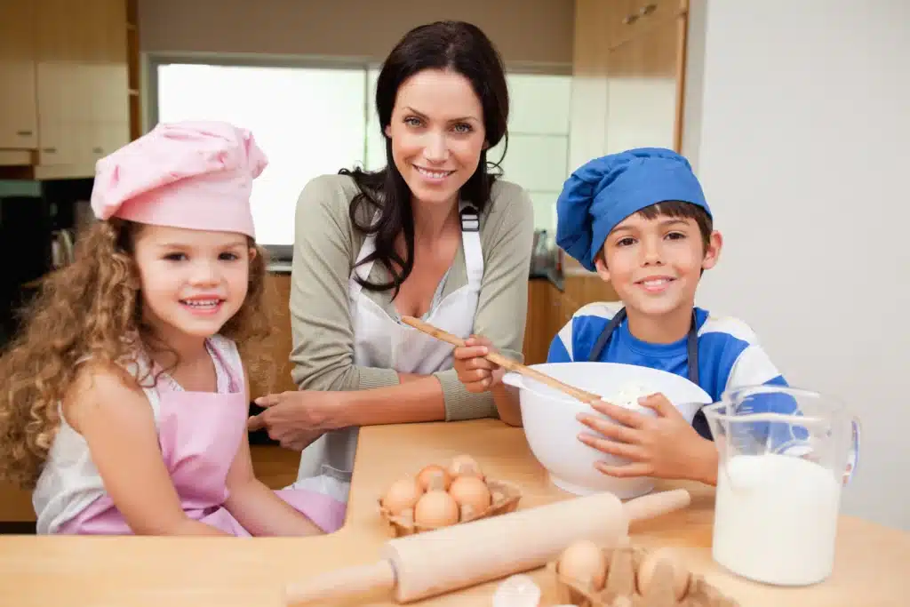 Home 1 A mother baking with her two children in the kitchen, smiling and mixing ingredients with eggs, milk, and baking tools on the counter.