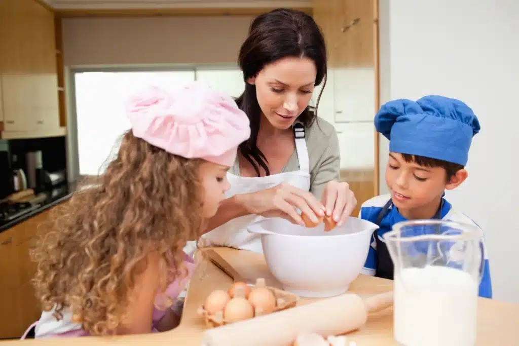 About Me 1 Olivia cracking an egg into a mixing bowl while her daughter in a pink chef hat and son in a blue chef hat watch attentively.